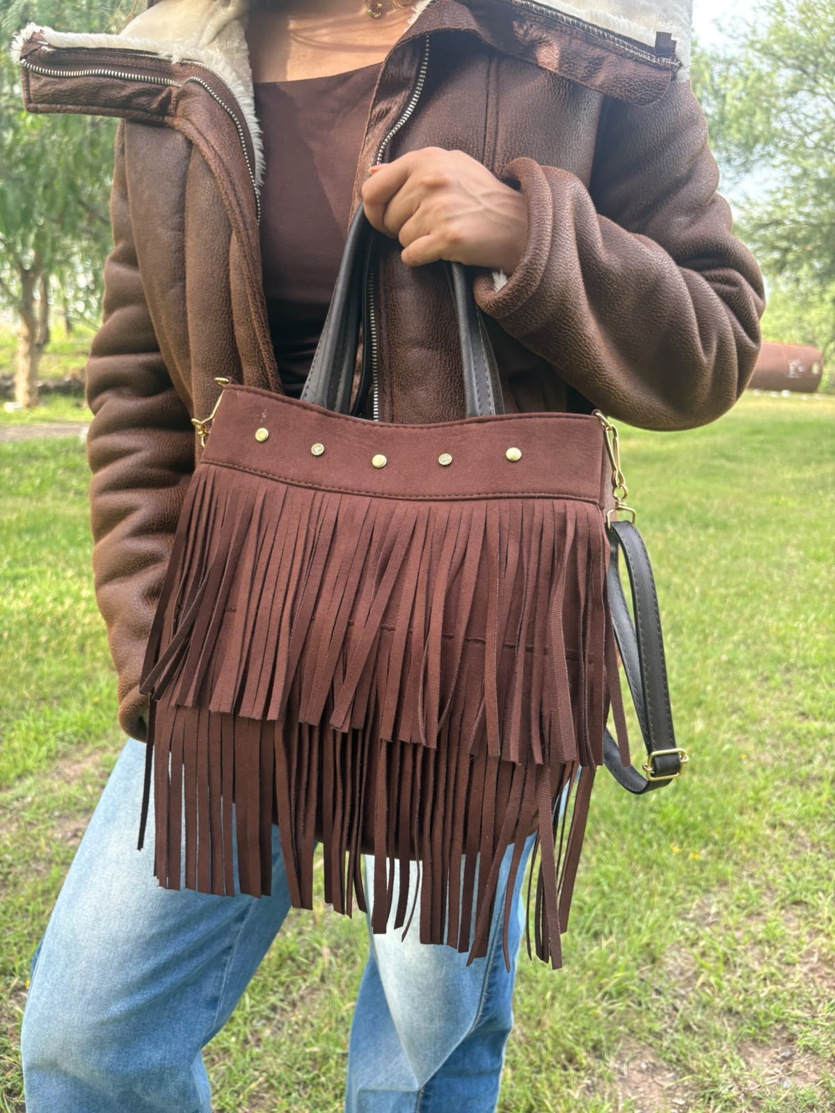 Bolso RAVENA de flecos en gamuza café, sostenido en la mano por una modelo en exterior